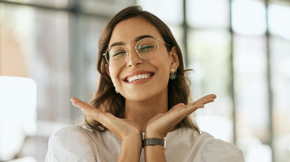 Junge Frau mit Brille umrahmt ihr Gesicht mit ihren Händen und grinst.