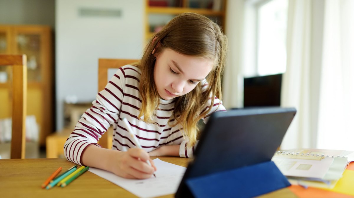 Mädchen, ca. 9 Jahre alt, sitzt mit einem Tablet, Papier und Stiften am Tisch und erledigt Aufgaben.