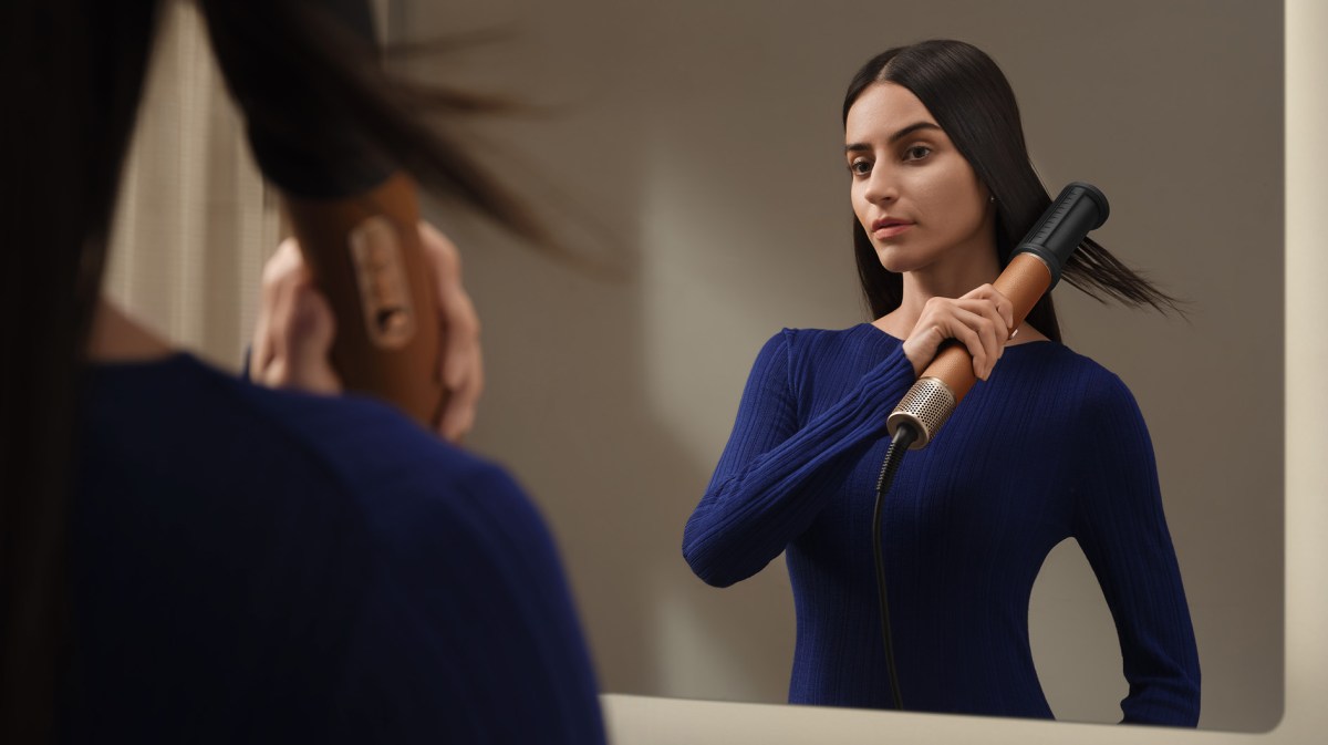 Brünette Frau im blauen Pullover glättet ihr Haar mit einem Haarstylinggerät vor einem Spiegel, ihr Blick ist fokussiert.