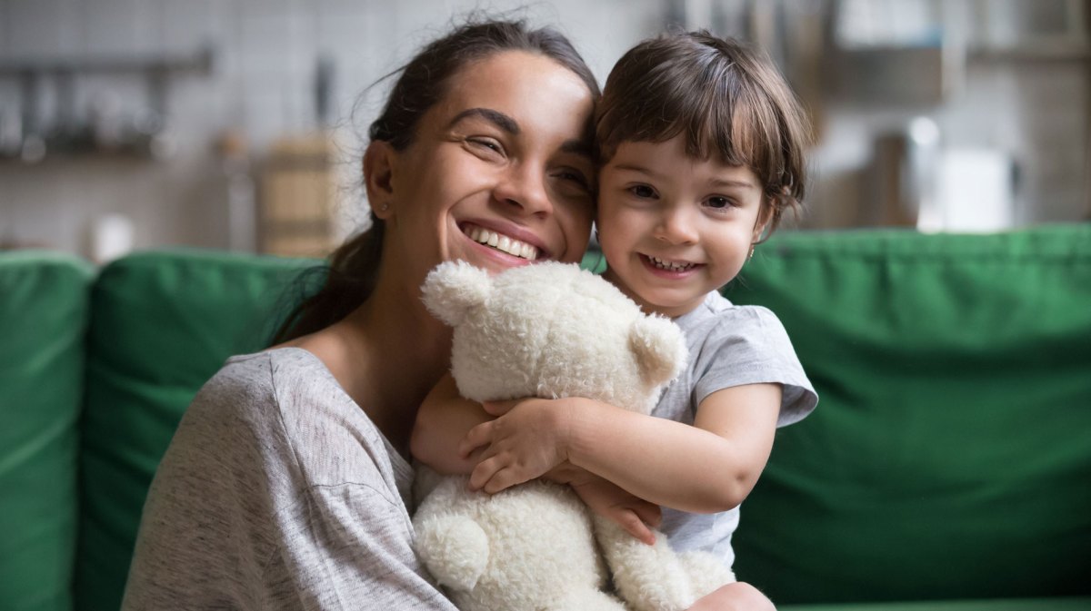 Mutter umarmt ihre Tochter, die einen Teddybären in den Armen hält. Beide sitzen im Wohnzimmer auf einer Couch.