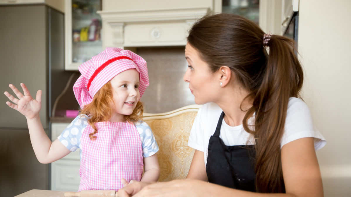 Mutter redet mit ihrer Tochter am Küchentisch, die eine rosa Kochmütze und Schürze an hat.