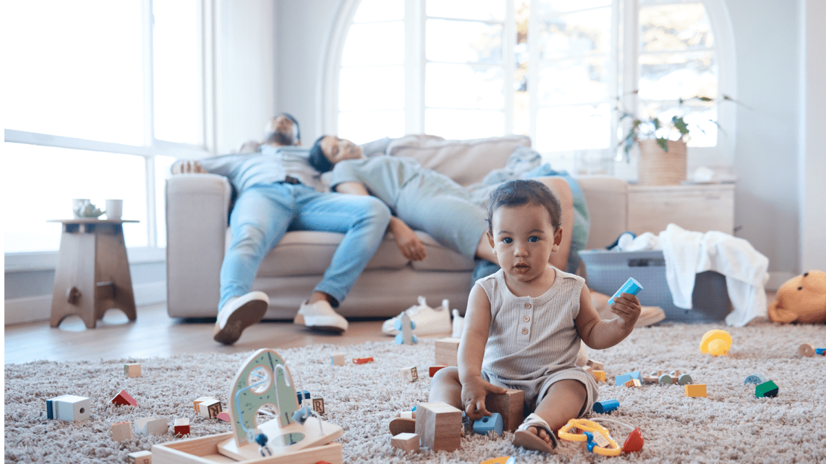 Schlafende Eltern liegen auf dem Sofa. Davor sind viele Spielsachen und ein Kleinkind