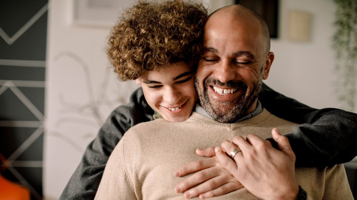 Porträt von Sohn und Vater. Der Sohn umarmt seinen sitzenden Vater von hinten. Beide lächeln zufrieden und glücklich.