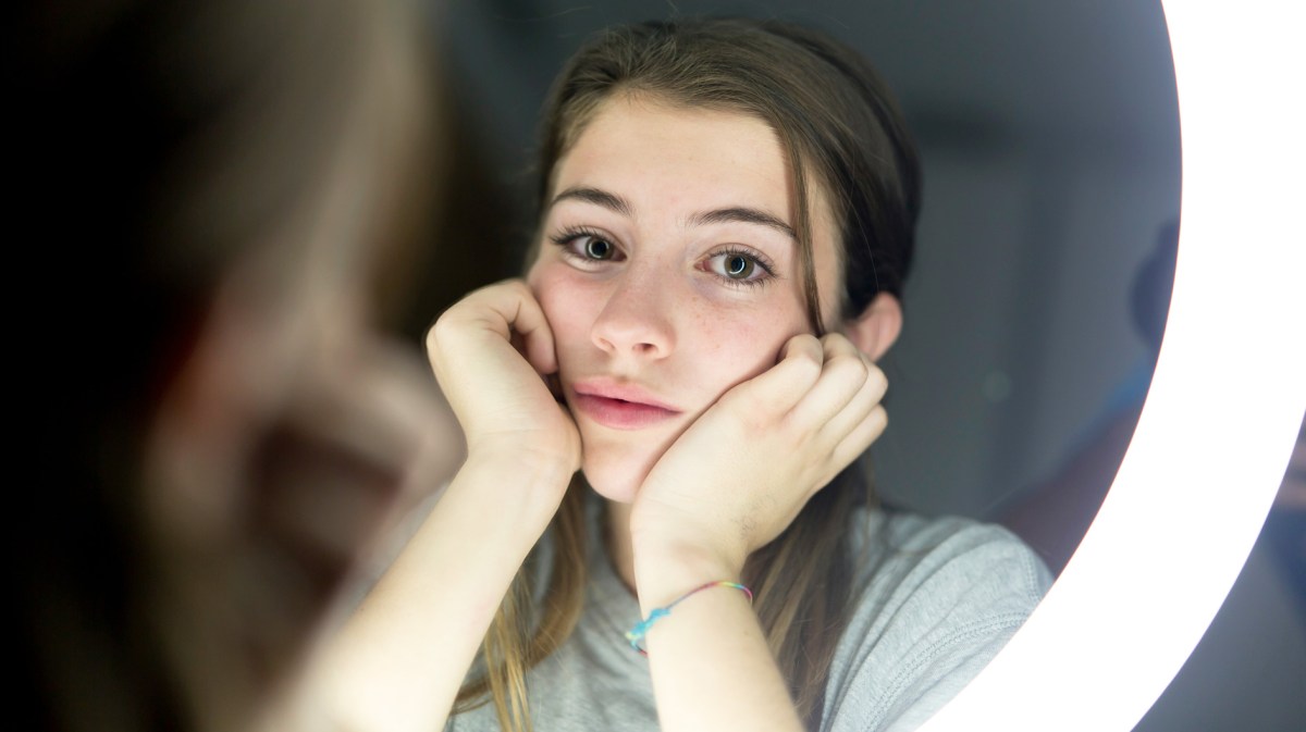 Porträt eines Mädchens im Teenageralter. Man sieht sein Bild durch einen Spiegel mit Ringlicht. Es hat den Kopf in beide Hände gestützt und sieht enttäuscht aus.