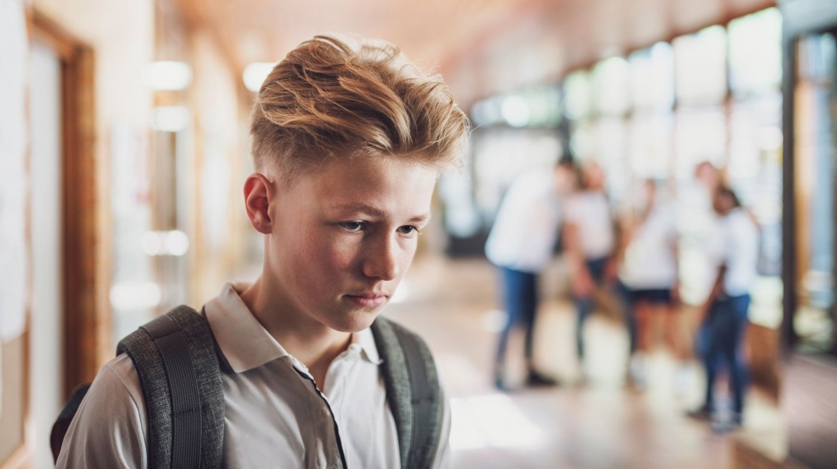 Männlicher Schüler, ca. 13 Jahre, geht mir gesenktem Kopf durch die Schule, im Hintergrund sieht man verschwommen andere Schüler, die scheinbar schlecht über ihn reden.