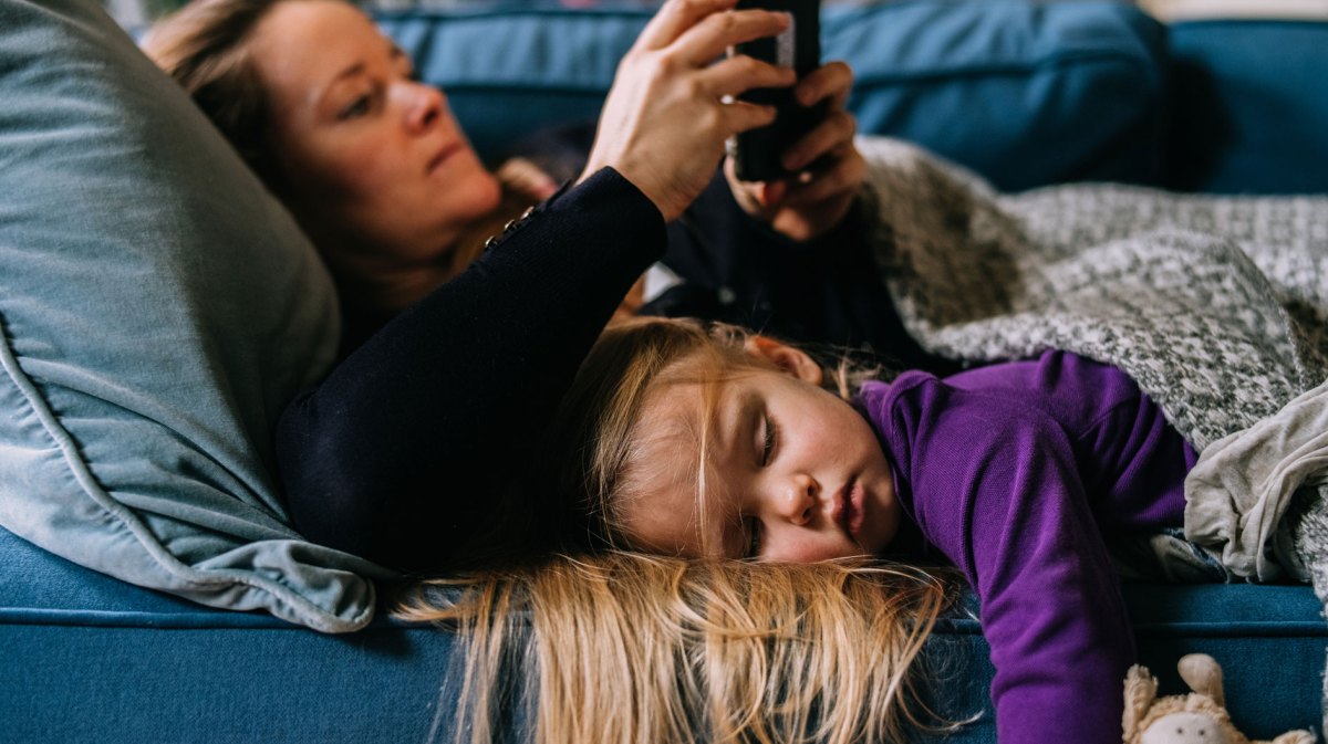 Mutter und Tochter liegen zugedeckt auf dem Sofa. Die Mutter schaut etwas auf ihrem Handy, während die Tochter im Vordergrund auf dem Bauch schläft.