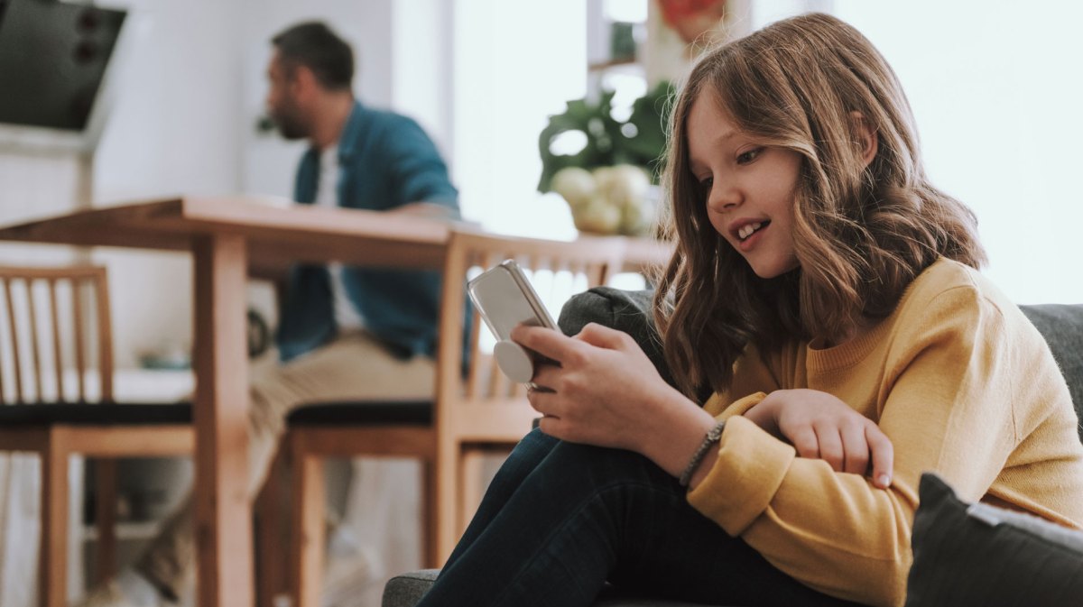 Mädchen, ca. 10 Jahre alt, sitzt mit ihrem Handy auf dem Sofa im Wohnzimmer. IM Hintergrund sieht man den Vater am Tisch sitzen.