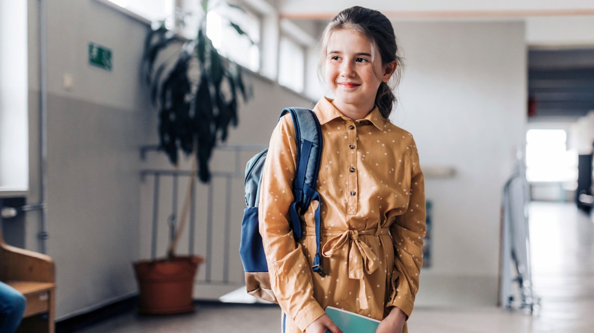 Mädchen, ca. 11 Jahre alt, steht in einem Schulgebäude, den Rucksack über einer Schulter gelegt, in den Händen ein Buch haltend.