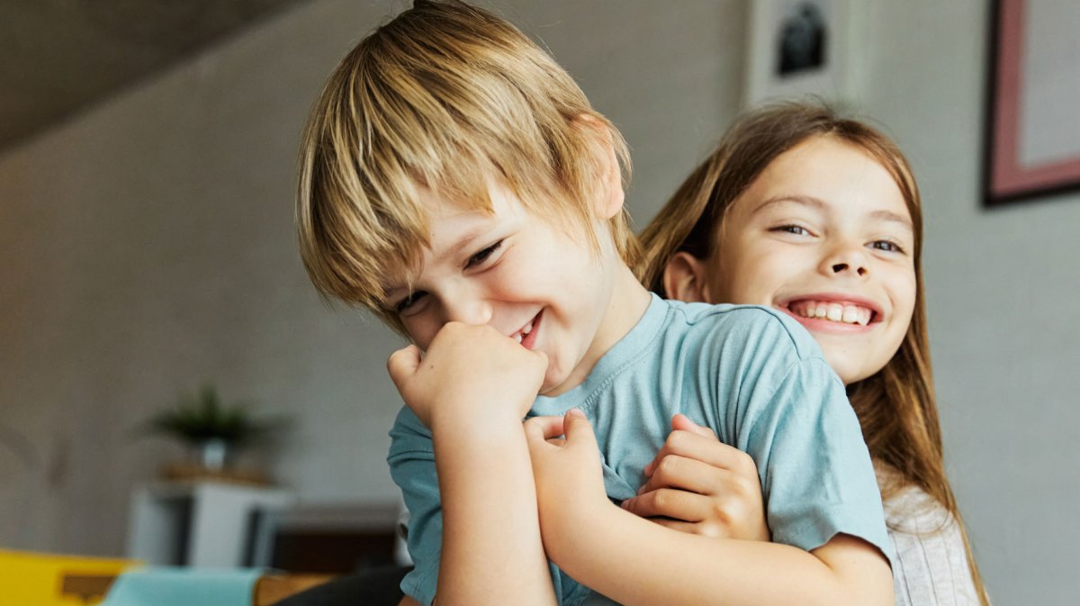 Kleiner Bruder sitzt auf dem Schoß der großen Schwester. Beide lachen und albern herum.