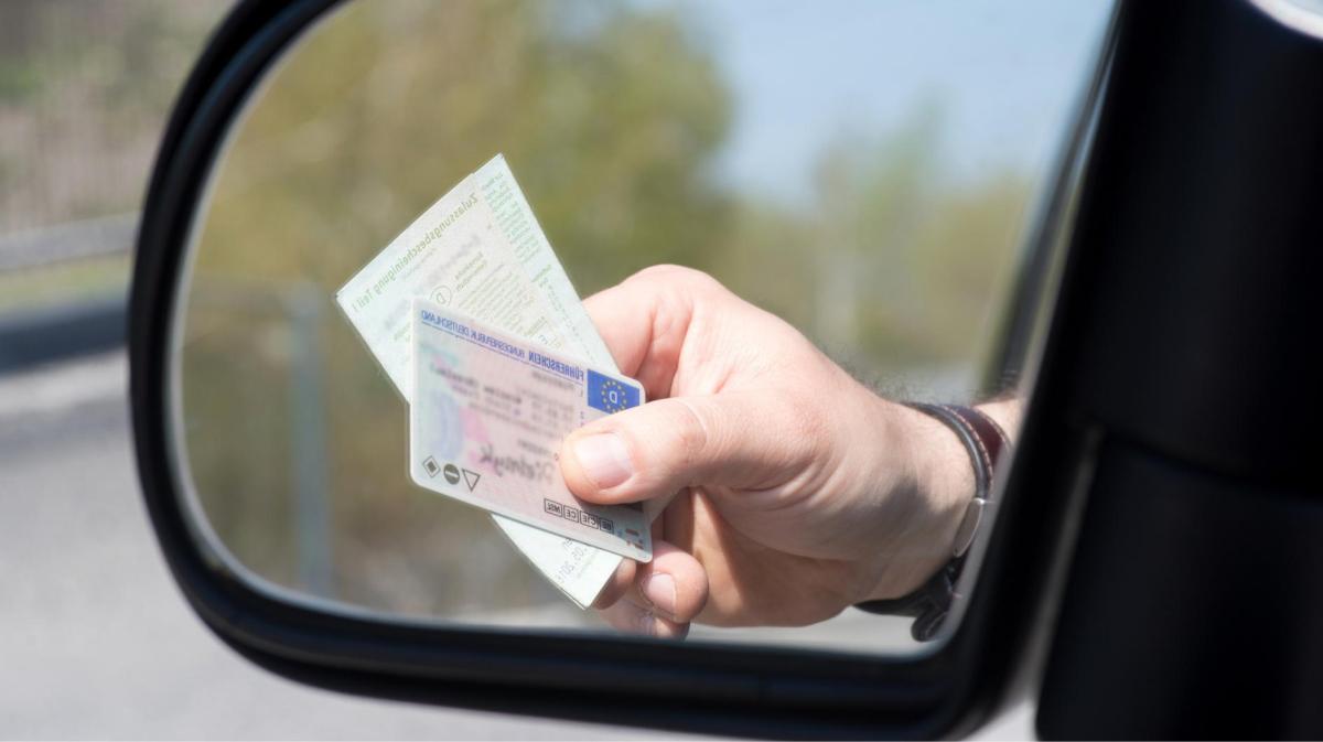 Person hält einen Führerschein und ein Ausweisdokument aus dem Auto, die im Seitenspiegel zu sehen sind.