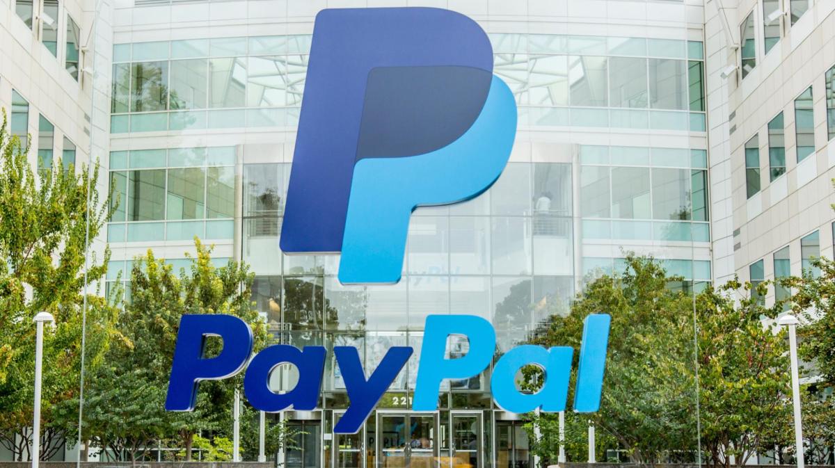 Das große PayPal-Logo in Blau vor dem modernen, verglasten Firmensitz mit weißen Fassaden und grünen Bäumen im Eingangsbereich.