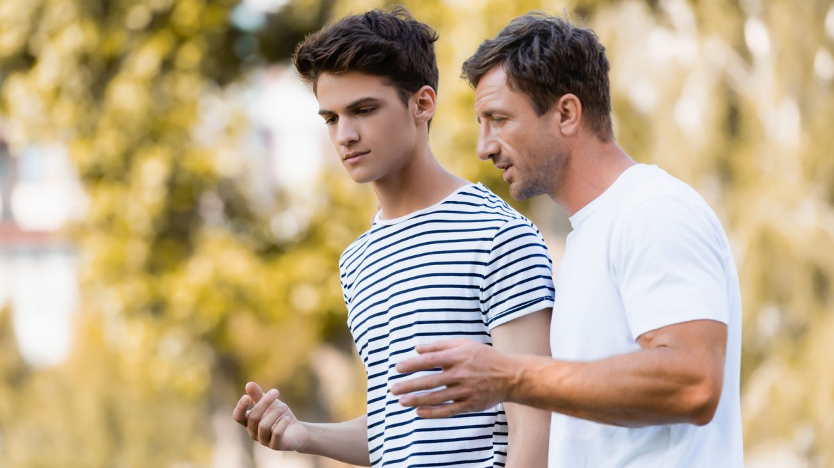 Vater und Sohn, ca. 16 Jahre alt, gehen an einem warmen Tag im Park spazieren und unterhalten sich ernst, aber freundlich miteinander.