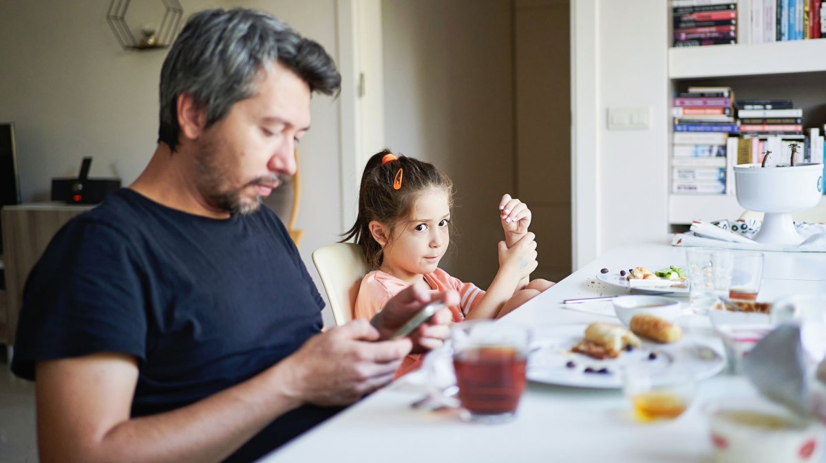 Vater und Tochter sitzen am Frühstückstisch. Der Vater schaut währenddessen auf sein Smartphone.